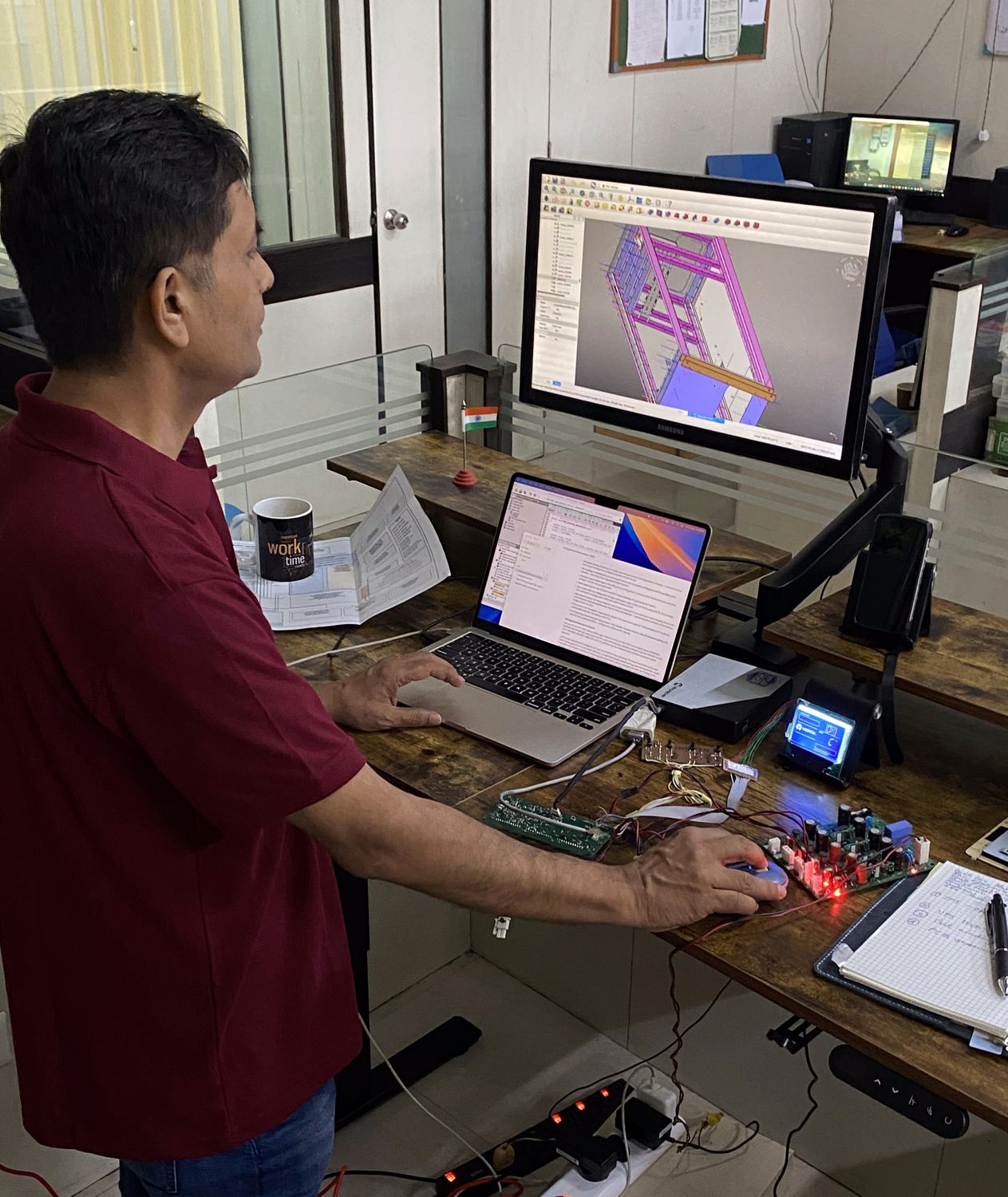 Engineer at a desk with a 3D CAD model of an enclosure on screen and a control board on the bench
