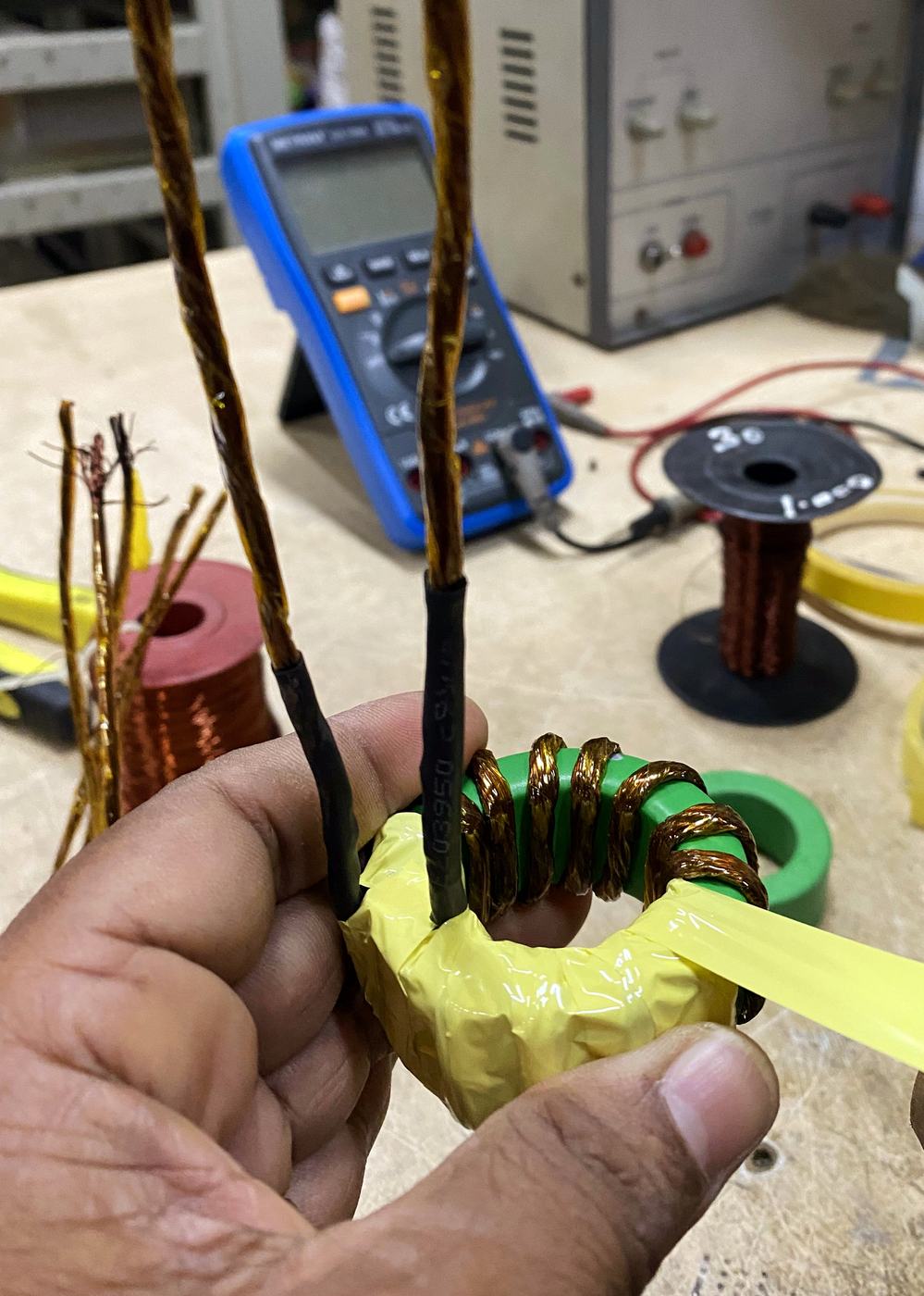 Hand holding a green toroidal core freshly wound with copper wire and yellow insulating tape, multimeter behind